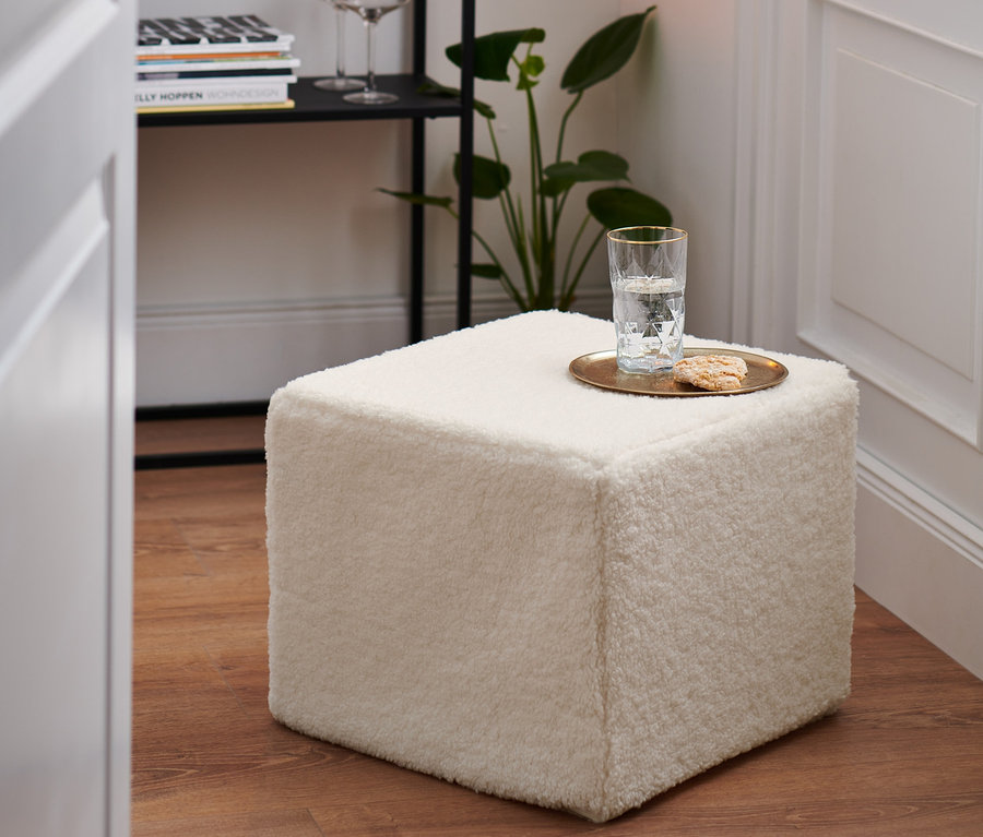 Un pouf crème est posé sur un plancher en bois. Un verre d'eau et un biscuit sont posés sur le pouf.