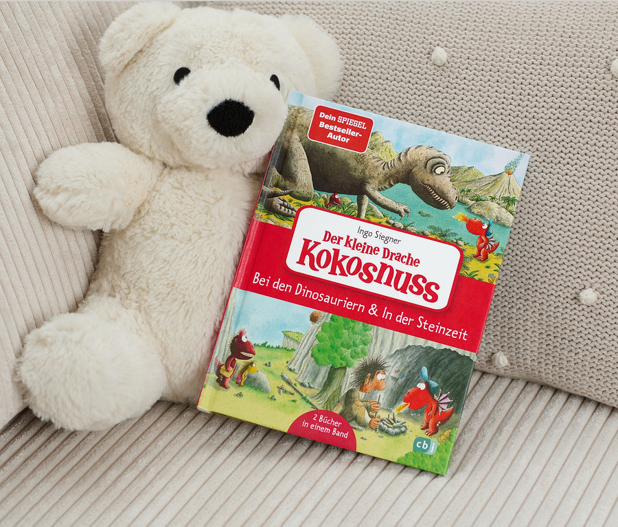 Un ours en peluche est posé à côté du livre « Der kleine Drache Kokosnuss – Bei den Dinosauriern & In der Steinzeit » (Le petit dragon Noix de coco – Chez les dinosaures et à l’âge de pierre, en allemand).