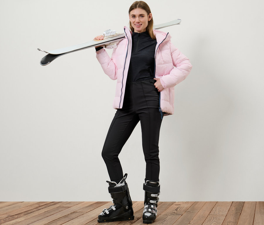 Une femme pose en legging de ski en softshell et chaussures de ski avec des skis sur l'épaule.