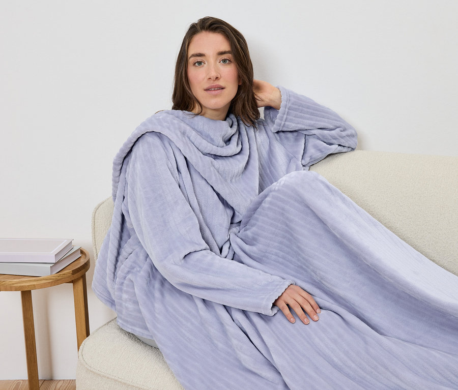 Une femme est assise sur un canapé enveloppée dans une couverture à manches lilas clair.