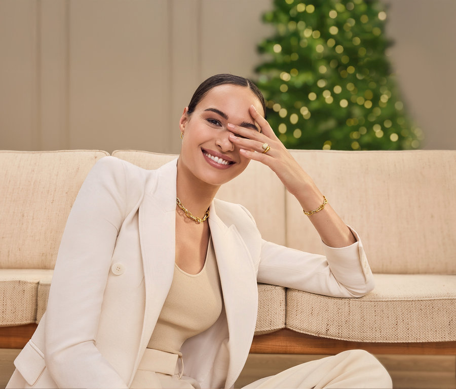 Une femme est assise sur un canapé, souriant et se couvrant un œil avec sa main. Elle porte une veste, un collier et un bracelet.