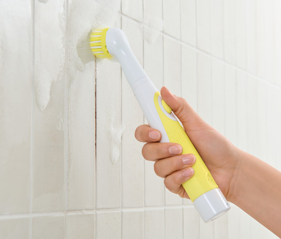 Une main tient une brosse de nettoyage électrique avec un manche jaune et nettoie des carreaux blancs