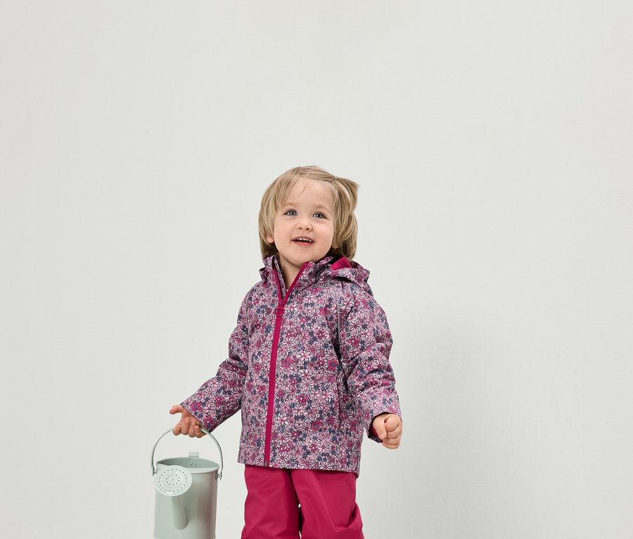 Un jeune enfant tient un petit arrosoir vert. Il porte une veste à fleurs et un pantalon rose.