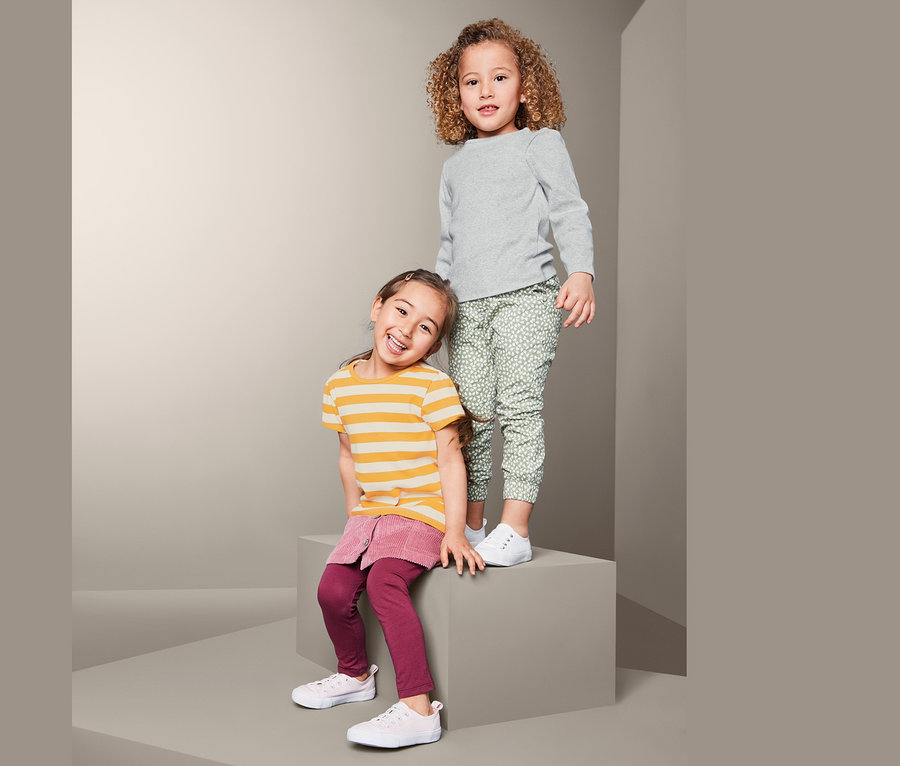 Deux filles posent sur un fond gris. L'une est debout et porte 2 t-shirts en matière côtelée gris et un pantalon vert imprimé. L'autre est assise et porte un t-shirt rayé jaune et une jupe rose.