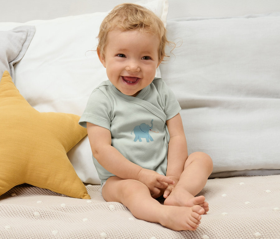 Un bébé souriant est assis sur un lit et porte un des 5 bodys évolutifs pour bébé avec un motif d'éléphant.