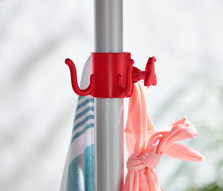 Sur un mât de parasol argenté sont fixés 2 crochets à parasol rouges auxquels sont suspendus une serviette et des vêtements roses.