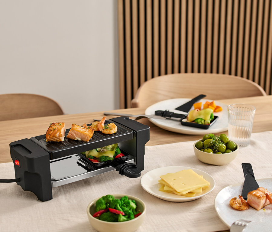 Un appareil à raclette pour 2 personnes est posé sur une table, avec du saumon et des crevettes sur le grill. Des assiettes d'ingrédients entourent le grill.