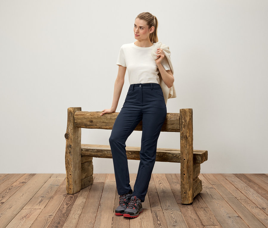Une femme appuie contre un banc en bois, un pull crème jeté sur l'épaule, portant un t-shirt blanc, un pantalon bleu marine et des chaussures de randonnée.