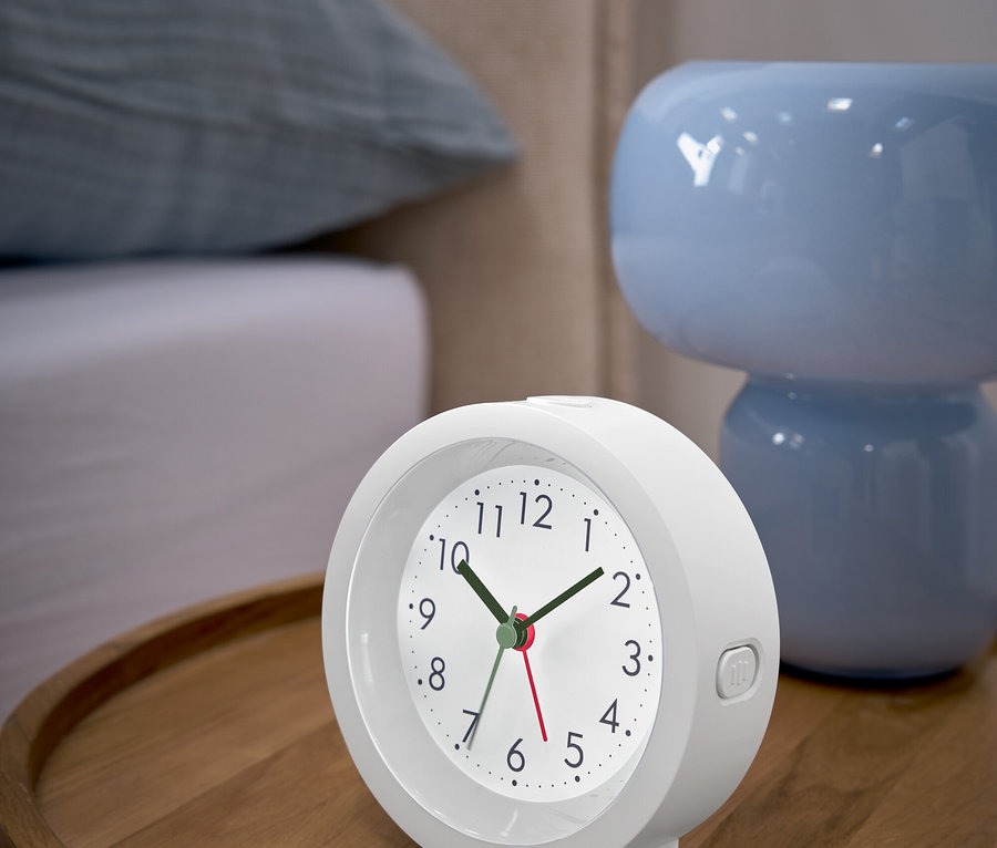 Un réveil blanc posé sur une table de chevet en bois à côté d'une lampe bleue.