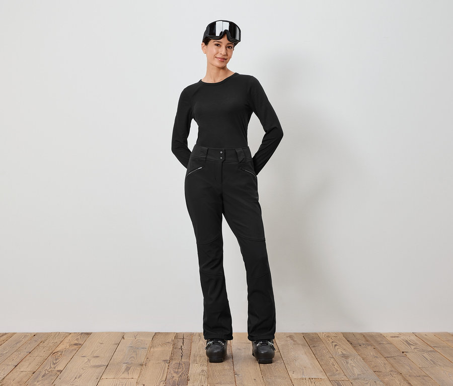 Une femme vêtue d'une tenue de ski noire et de lunettes de ski sur la tête se tient devant un mur blanc.