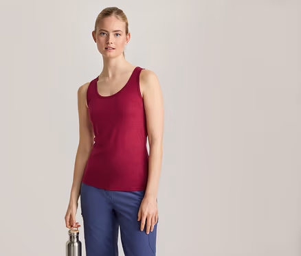Une femme aux cheveux blonds attachés, vêtue d'un débardeur magenta et d'un pantalon bleu, se tient de face et tient une bouteille d'eau argentée dans sa main droite.