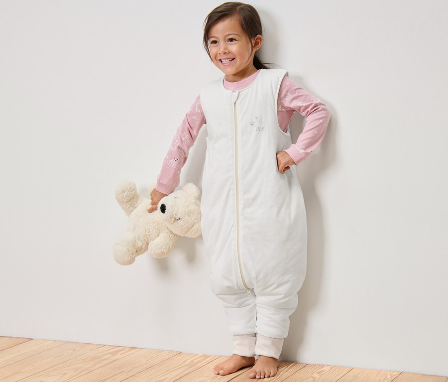 Une fillette souriante debout tenant un ours en peluche, portant une gigoteuse avec jambes, 2,5 TOG, 110 cm.