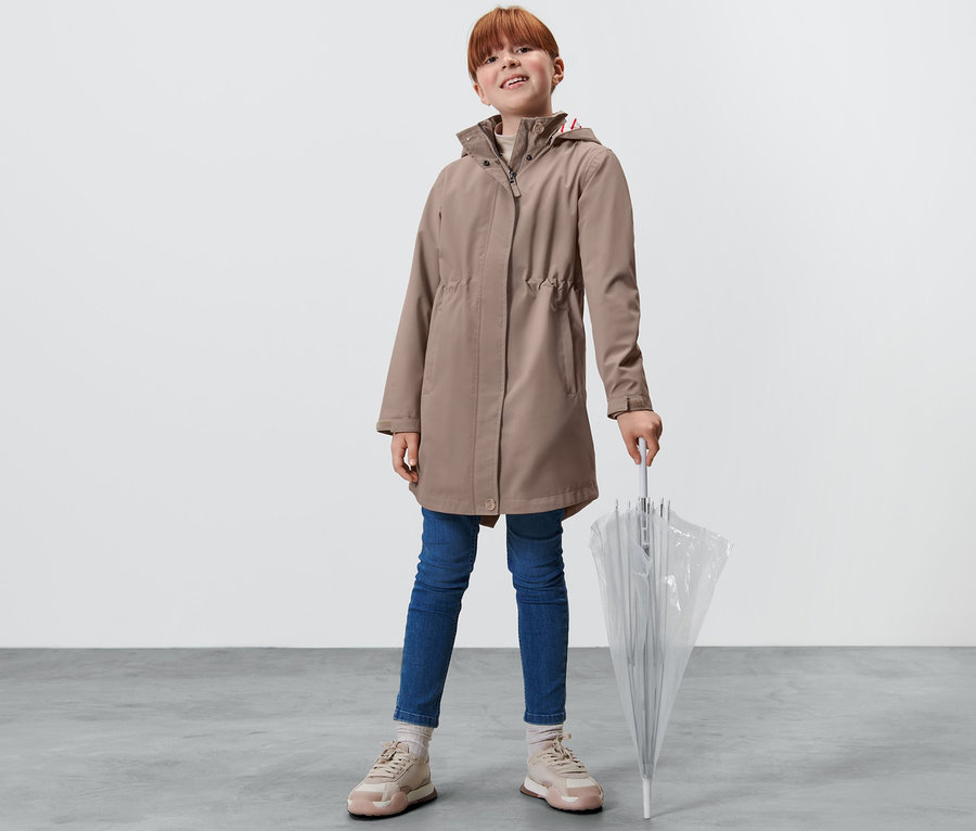 Une fille debout tenant un parapluie transparent. Elle porte un trench-coat tout-temps pour enfant, un jean et des baskets.