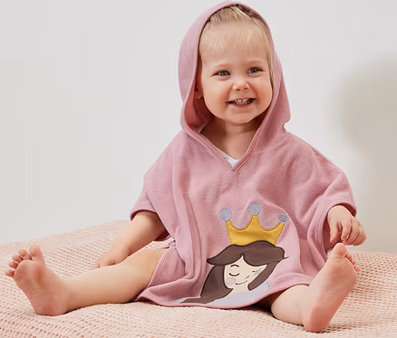 Un enfant souriant est assis sur une surface rose et porte un poncho en éponge à capuche avec un motif de princesse.