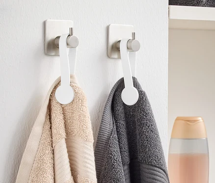 Deux serviettes, une beige et une grise, sont suspendues à des crochets blancs sur le mur. À l'arrière-plan, on peut voir des serviettes grises pliées et une bouteille.