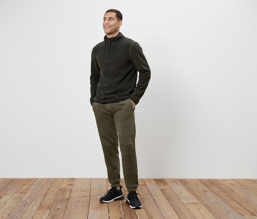 Un homme portant un pantalon vert olive et un pull en polaire gris foncé à fermeture éclair, debout sur un plancher en bois.