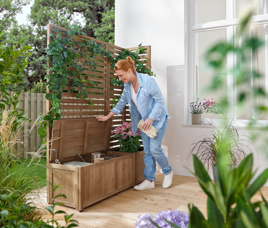 Une femme ouvre un banc de jardin en bois avec rangement, à côté d'une clôture en bois avec des plantes grimpantes.