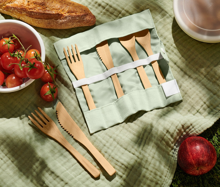 Sur une couverture verte sont disposés du pain, des tomates, des couverts de pique-nique, une pomme et un récipient.