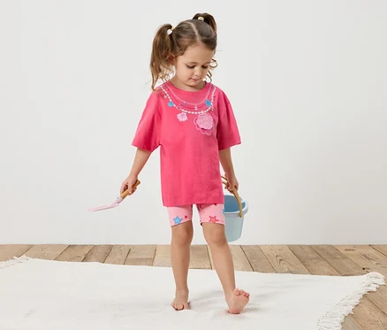 Une petite fille avec des tresses se tient pieds nus sur un tapis blanc. Elle porte un t-shirt rose avec un décor de coquillages et un short rose avec des étoiles. Dans ses mains, elle tient une pelle et un seau bleu.