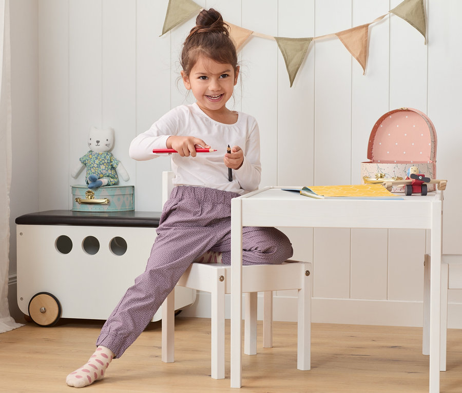 Fillette assise à une table tenant des crayons. Elle porte un pantalon à enfiler en coton.