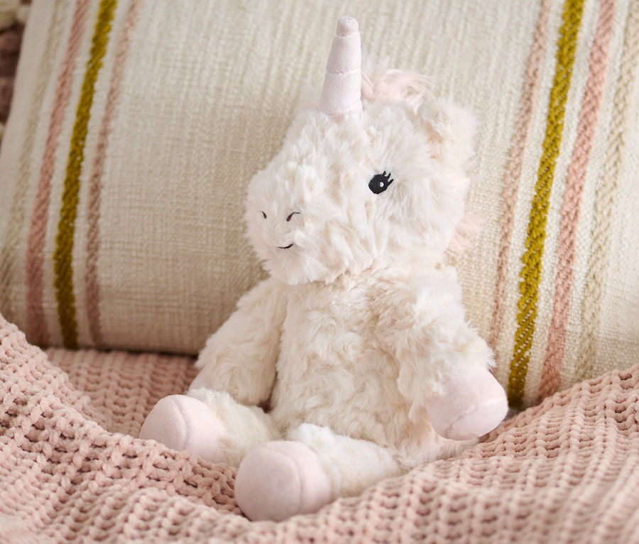 Une peluche « Licorne » est assise sur une couverture en tricot rose à côté d'un coussin beige.