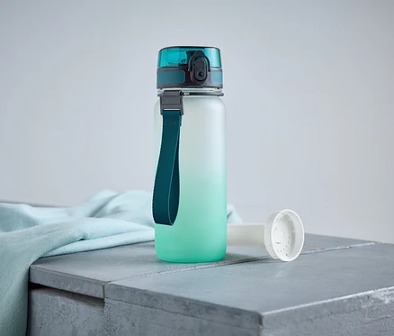 Une bouteille d'eau avec un bouchon et une lanière turquoise est posée sur une surface grise à côté d'un tissu bleu clair.