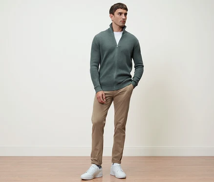 Un homme debout portant une jaquette en maille sauge et un pantalon chino sable.