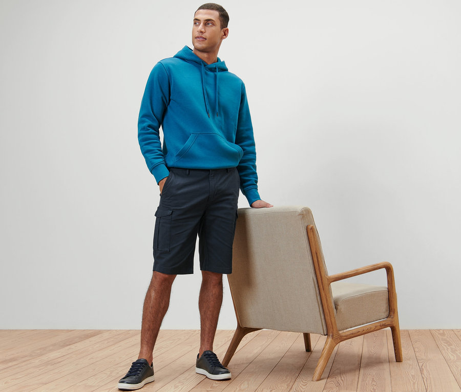 Un homme pose en sweat à capuche bleu et short cargo bleu marine.