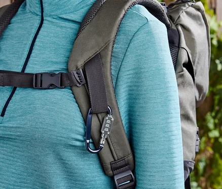 Gros plan d'une femme portant un sac à dos vert olive et un haut turquoise devant un fond de clôture en bois et de plantes.