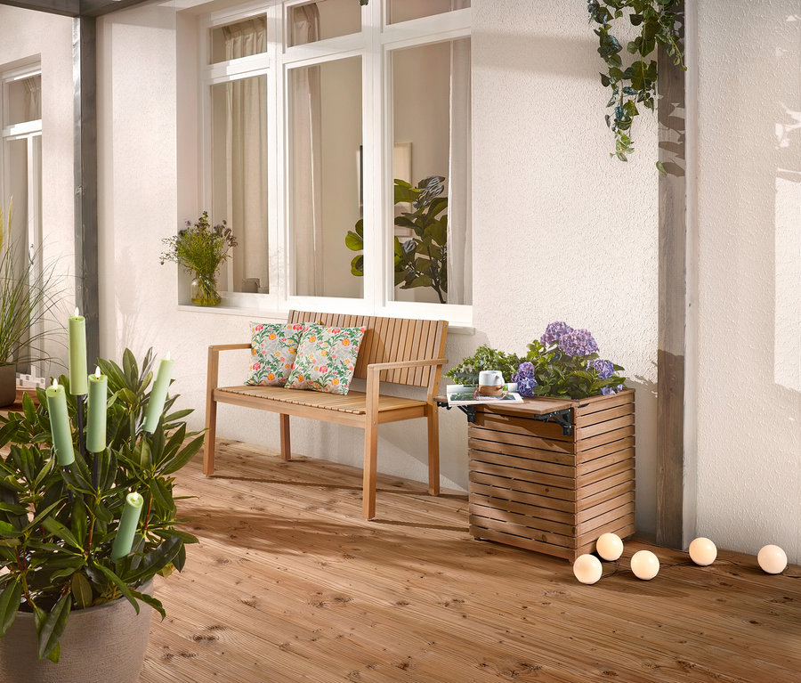Sur une terrasse en bois se trouvent un banc en bois avec des coussins, un bac à plantes avec des fleurs et une plante en pot avec des bougies.