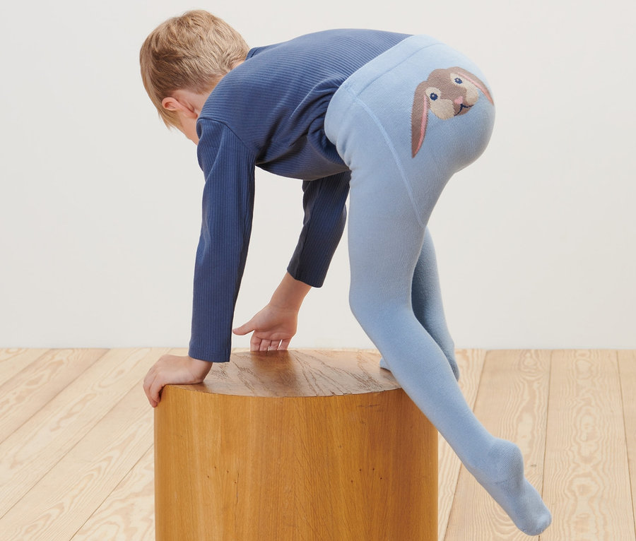 Un enfant s'appuie sur un tabouret en bois, il porte un collant bleu avec un dessin de lapin sur les fesses. Un jouet en peluche est posé sur le sol. 2 collants pour bébé et tout-petit.