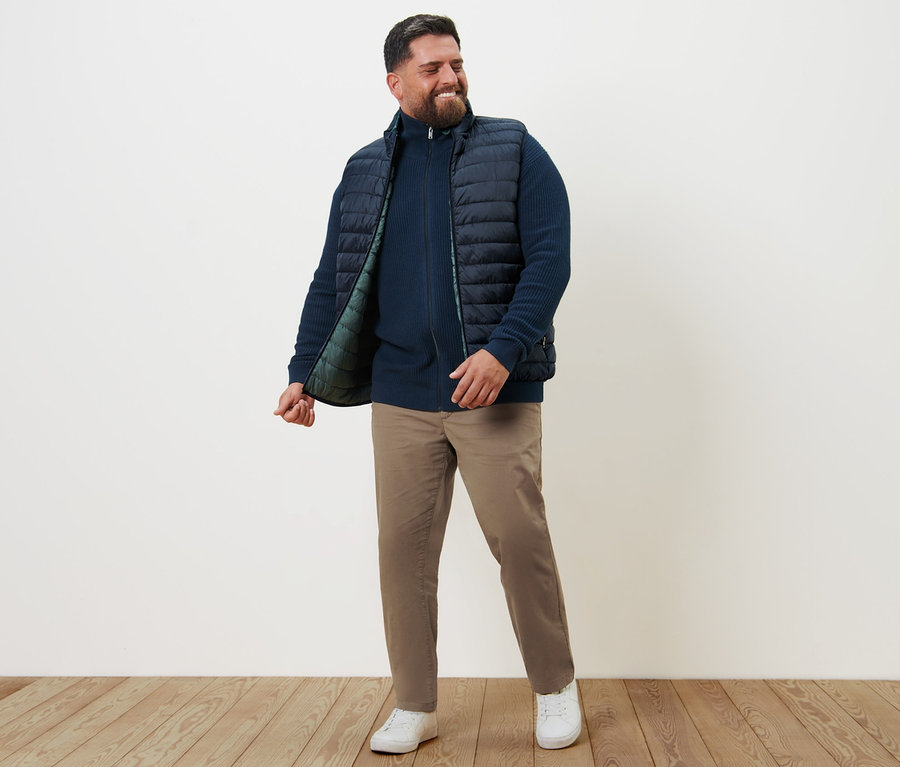 Un homme souriant se tient debout et porte un gilet réversible, une jaquette en maille bleu marine et un pantalon chino sable.
