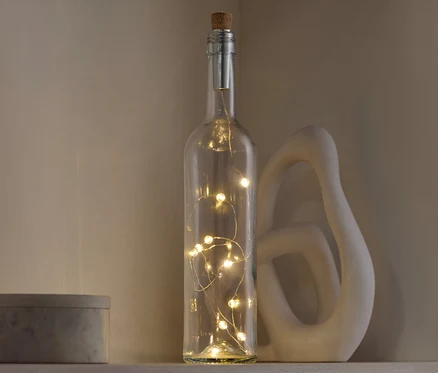 Une bouteille décorative en verre avec un bouchon en liège et des lumières à l'intérieur, posée sur une étagère à côté d'une sculpture décorative et d'une boîte grise.