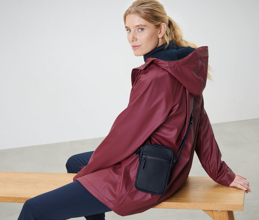 Une femme blonde est assise sur un banc, portant une veste bordeaux et un sac à bandoulière de pluie noir.