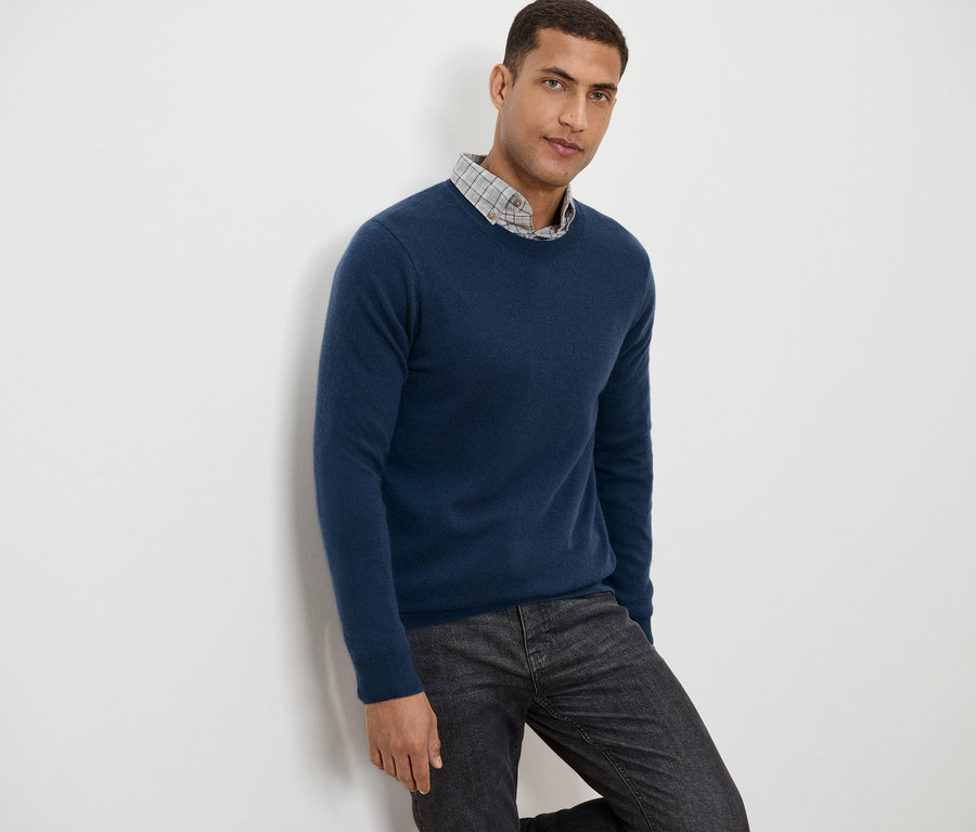 Un homme appuyé contre un mur portant un pull bleu et un pantalon gris à motifs.