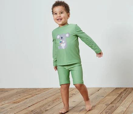 Un garçon portant une combinaison pour enfants avec protection UV 80, koala, se tient sur un plancher en bois à côté d'un jouet.