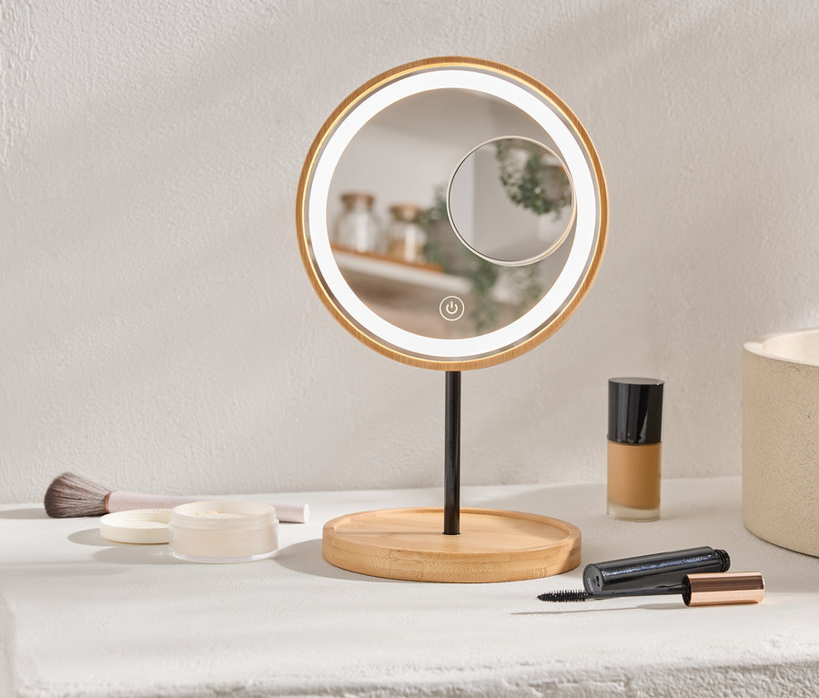 Sur une étagère se trouvent un miroir de maquillage rond et éclairé, du maquillage et un pinceau.