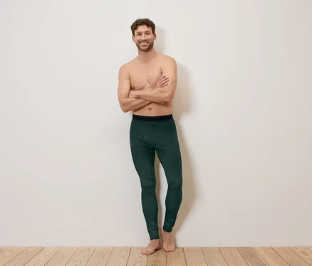 Un homme bras croisés, pieds nus, porte un legging vert foncé devant un mur blanc.