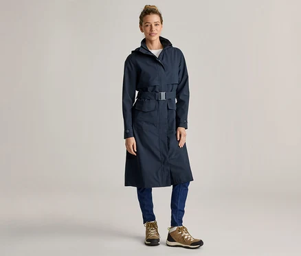 Une femme portant un chignon porte un long imperméable bleu marine ceinturé, un jean bleu et des chaussures de randonnée marron sur un fond gris clair.