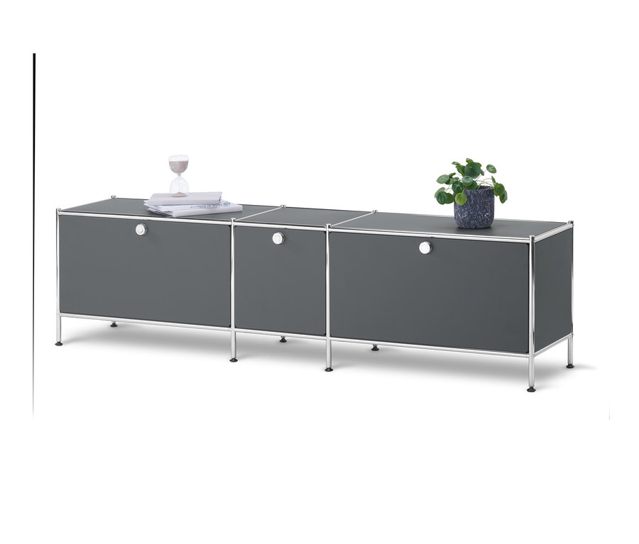 Un meuble bas gris « CN3 » avec 3 abattants sur un fond blanc. Sur le meuble bas se trouvent une plante en pot, des livres et un sablier.