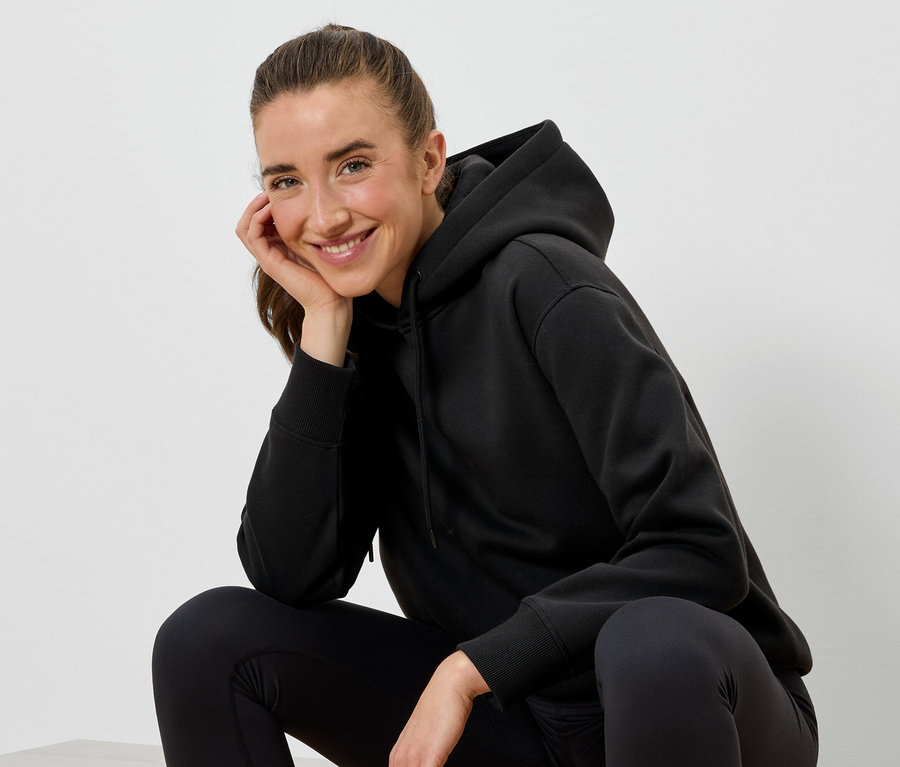 Une femme souriante portant un sweat à capuche noir et un legging noir est assise, le visage appuyé sur sa main.