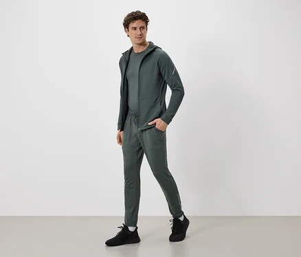 Homme en survêtement vert olive avec veste à capuche et pantalon à cordon de serrage, et baskets noires.