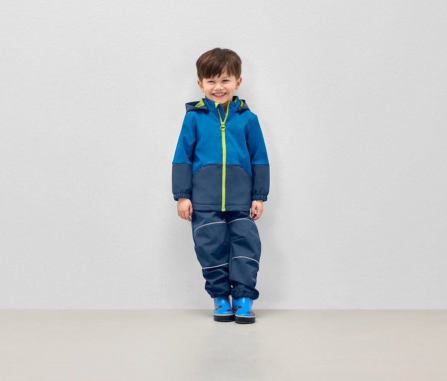 Garçon souriant posant en veste bleue à capuche et pantalon bleu marine avec bandes réfléchissantes.