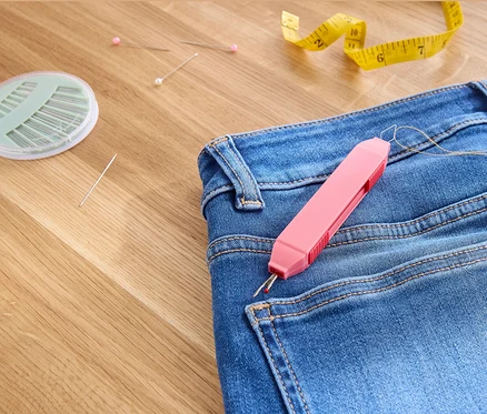 Jean avec un outil de couture 2-en-1, des aiguilles, des épingles et un mètre ruban jaune sur une surface en bois.