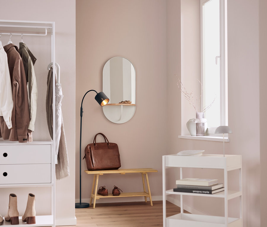 Dans un intérieur, on trouve une étagère blanche avec des vêtements, un miroir ovale, un sac en cuir marron sur un banc, une lampe noire et un chariot à roulettes blanc.