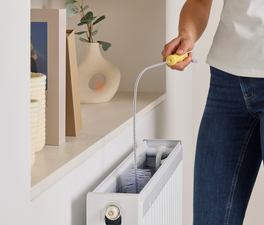 Une femme nettoie un radiateur blanc avec une brosse flexible. Des décorations sont sur l'étagère.