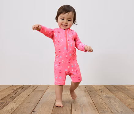 Un jeune enfant marche pieds nus sur un plancher en bois, vêtu d'un maillot de bain rose avec une fermeture éclair et un motif d'hippocampes, de coquillages et d'étoiles de mer.