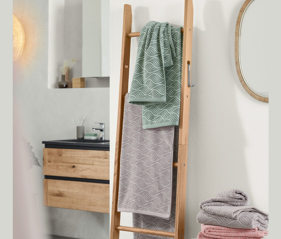 Dans la salle de bain, une échelle en chêne sur laquelle sont suspendus 2 linges de toilette en jacquard et un linge de bain en jacquard. À côté de l'échelle se trouve une pile de linge de bain et de toilette en jacquard.