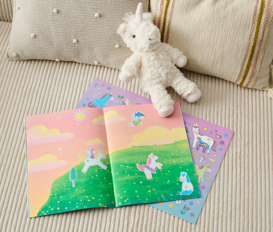 Sur un canapé sont posés un livre ouvert avec des illustrations de licornes, à côté d'une peluche de licorne et du livre d'autocollants « Mon livre d’autocollants métallisés « Le monde merveilleux des licornes » (en allemand) ».