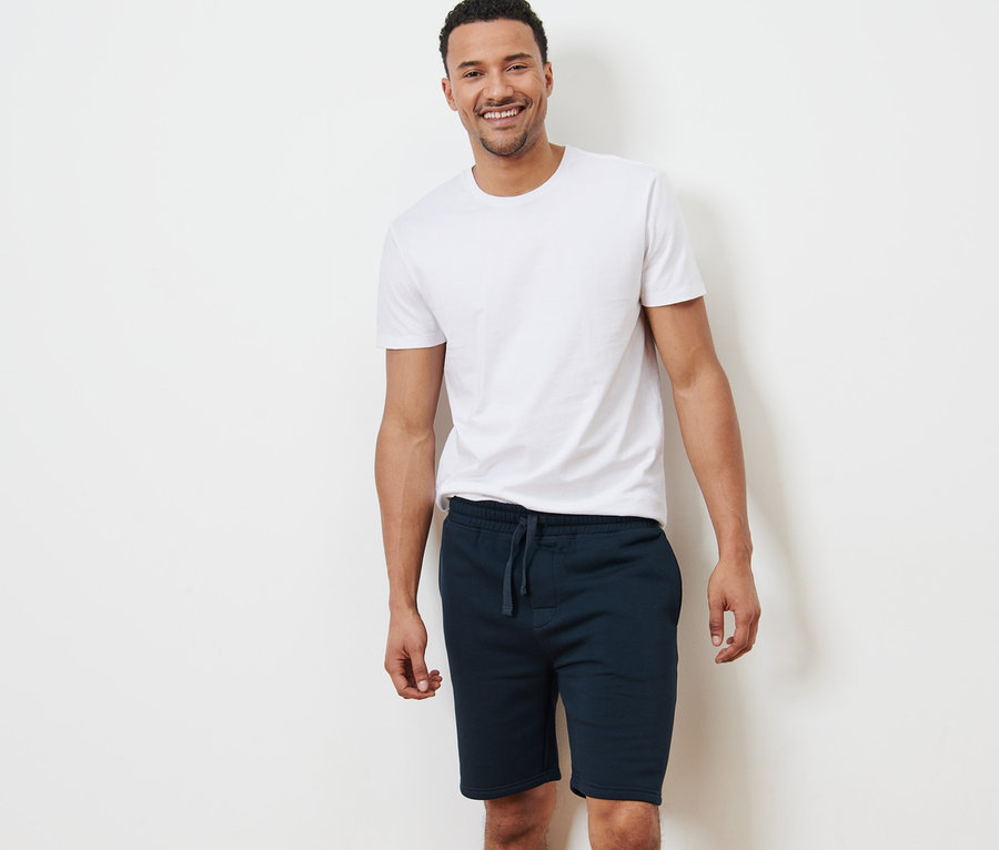 Un homme souriant se tient devant un mur blanc, portant un t-shirt blanc et un short en sweat.